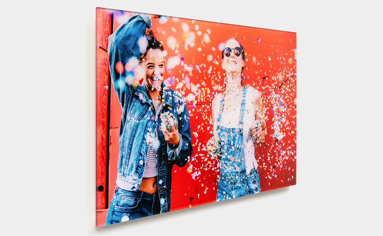 Angled shot of a 20x30" Acrylic Print featuring a photo of two women in front of a red wall throwing confetti. 