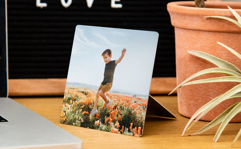 4x4" Metal Print with an Easel Back styled on a desk, the Metal Print has a photo of a young boy in a field of flowers on it. 
