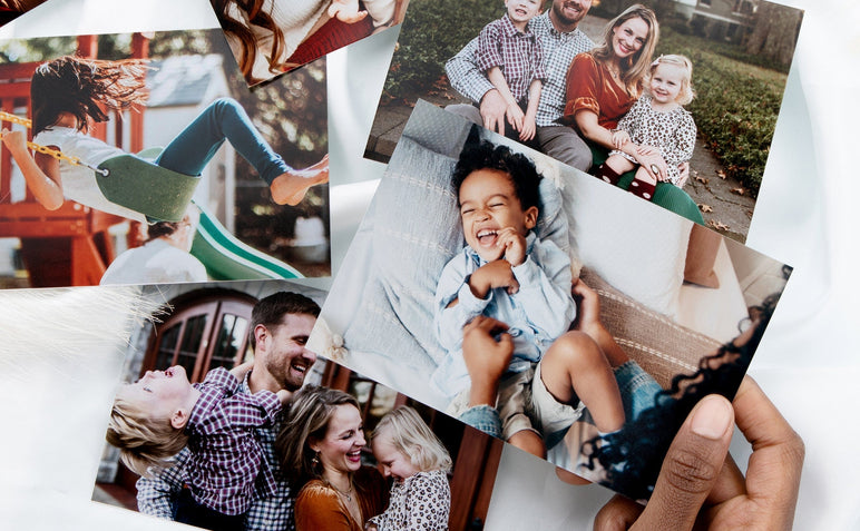 4x6" Photo Prints scattered on a white surface. The Prints feature families and child, one print with a laughing child is being held up by a woman's hand. 