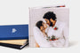 Three 8x8" Hardcover Photo Books with cloth covers stacked and another 8x8" Hardcover Photo Book with a lustre cover featuring a wedding photo standing in front of the stack. 
