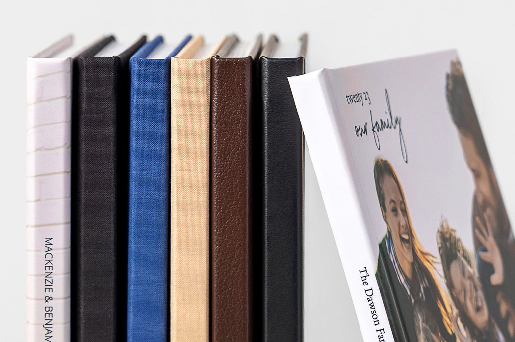 Seven Hardcover Photo Books display the different cover options: Lustre Photo Wrap, Black Cloth, Navy Cloth, Beige Cloth, Mahogany Leather, and Black Leather.  