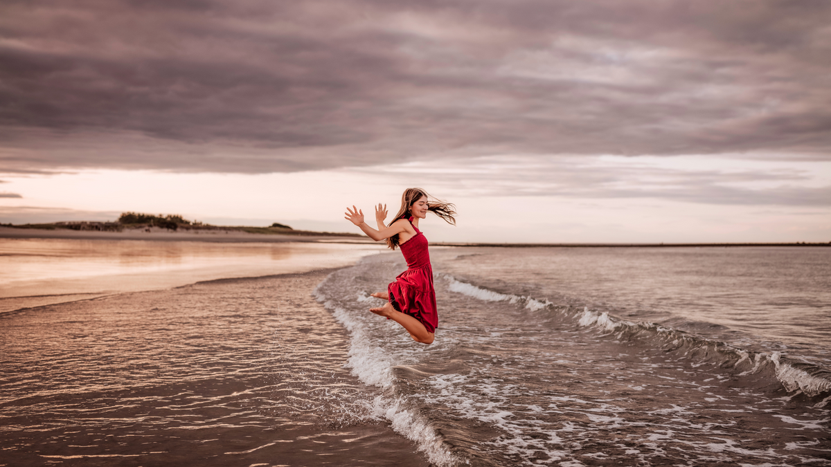 A girl in a bright red dress is captured mid-jump at the edge of the ocean, with her hair flowing and waves splashing, under a dramatic pink and purple evening sky.