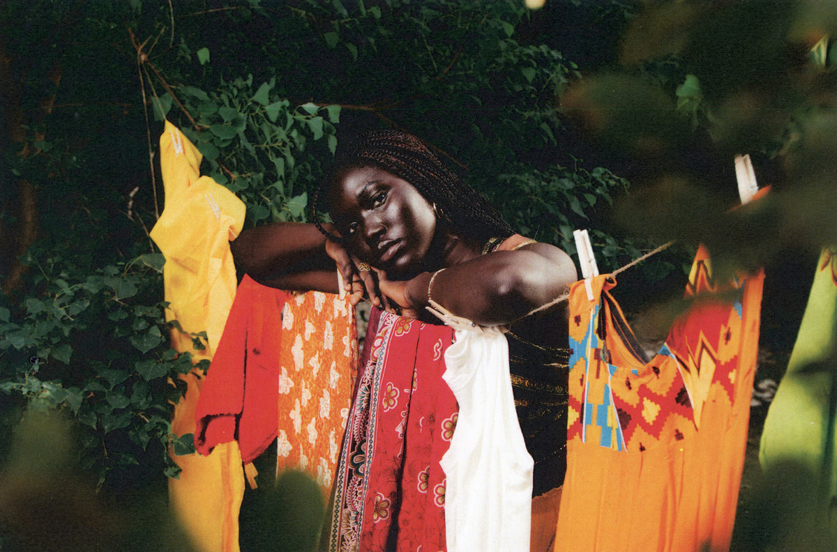 Tenny Rudolph's work: A woman rests her hand and arms on clothesline with bright orange,  yellow, and red clothing hanging. The portrait is frame by lush green foilage and trees,