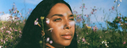 A woman standing in a field of wildflowers, looking away from the camera with a soft expression, framed by the flowers around her.