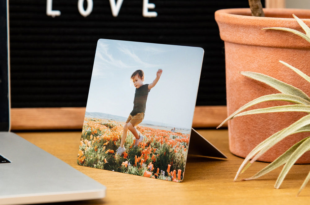 4x4" Metal Print with an Easel Back styled on a desk, the Metal Print has a photo of a young boy in a field of flowers on it. 