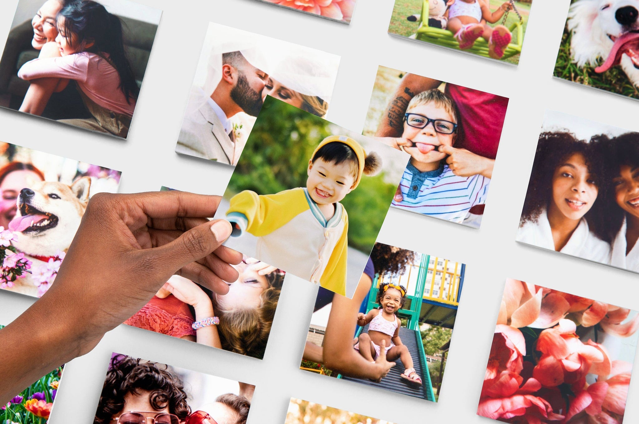 Flat lay of multiple square Photo Prints, the focus is a hand holding up a square Photo Print of a small child in a yellow hat.