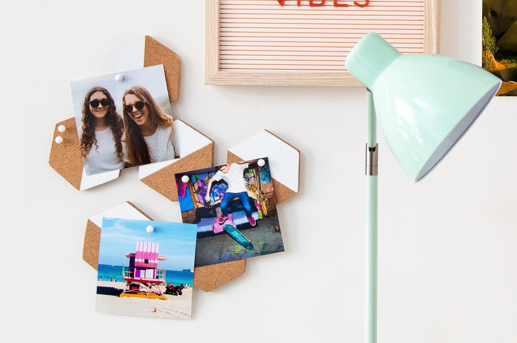 Lifestyle scene of three square Photo Prints tacked up on a wall next to a lamp and a letterboard. The Photo Prints feature pictures of teens and one photo of the beach.