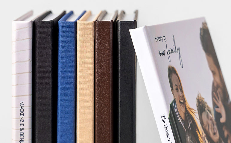 Seven Lay Flat Photo Books display the different cover options: Lustre Photo Wrap, Black Cloth, Navy Cloth, Beige Cloth, Mahogany Leather, and Black Leather.  